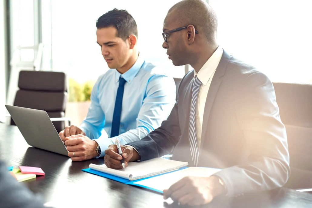 Two businessmen in suits collaborate at a conference table, reviewing a laptop while one takes notes in a notebook.