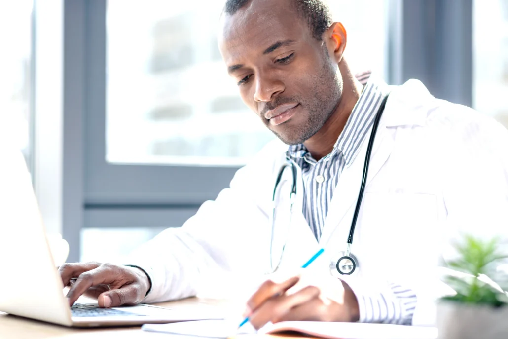 A male doctor wearing a white coat and stethoscope types on a laptop while taking notes in a notebook at a desk.