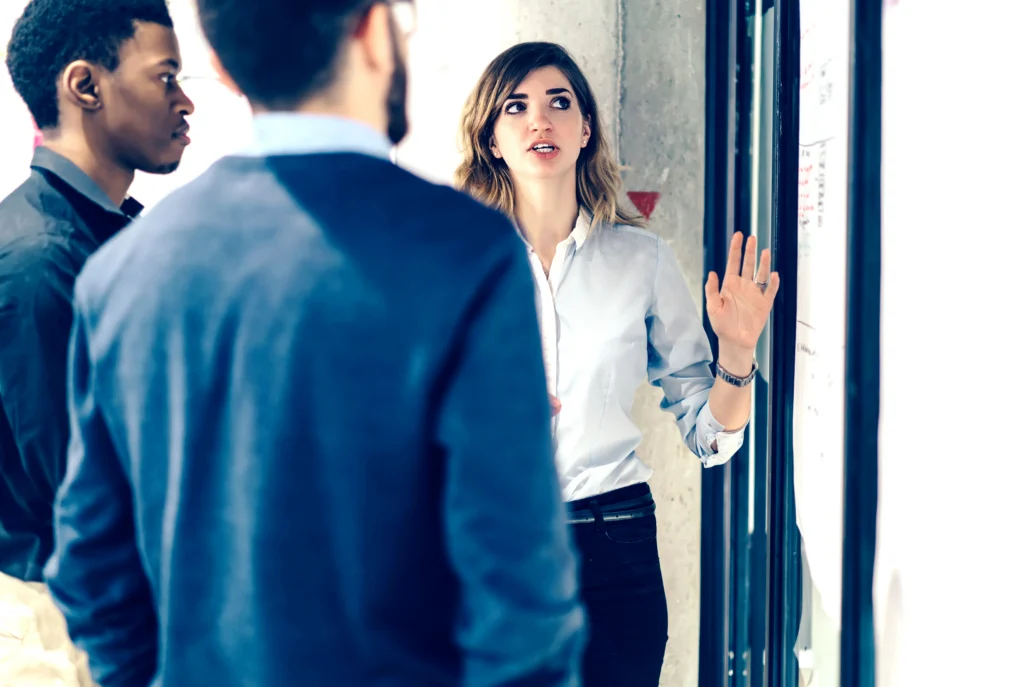 A woman in a white button-down shirt gestures while explaining something on a whiteboard to two attentive male colleagues in a modern office setting.