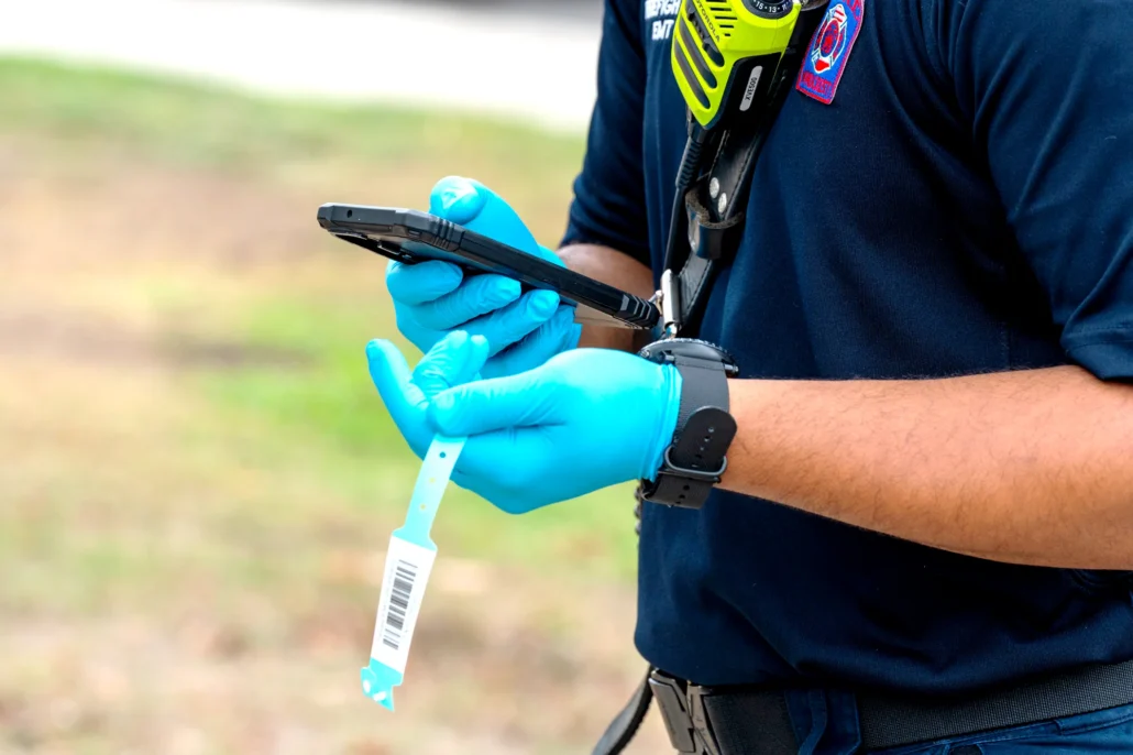 An emergency responder in blue gloves holds a smartphone and a patient ID wristband while wearing a radio and uniform patch outdoors.
