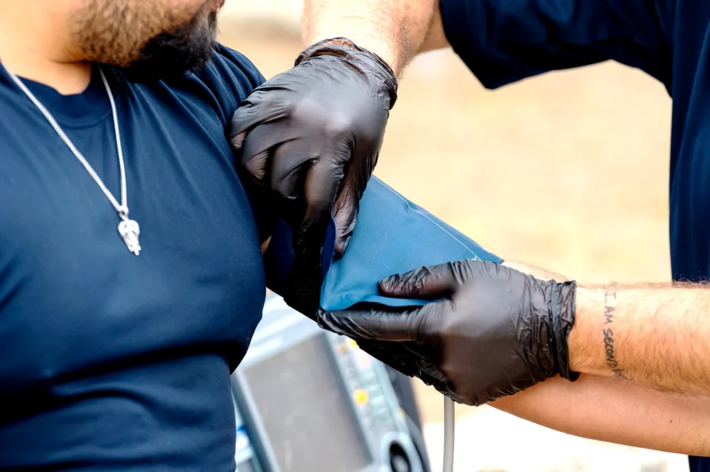 A healthcare worker in black gloves wraps a blue blood pressure cuff around a patient's arm outdoors.