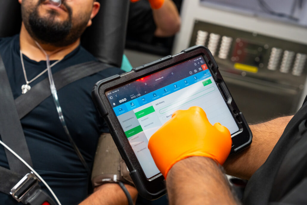 An emergency responder in orange gloves uses a rugged tablet to enter patient data while treating a man in an ambulance.