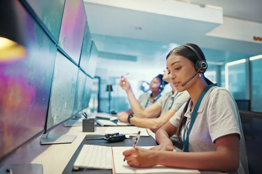 A woman wearing a headset sits at a desk with multiple monitors, taking notes while monitoring a control room.