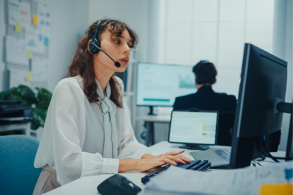 A customer service representative wearing a headset types on a keyboard while looking at the computer screen in a busy office with colleagues working in the background.