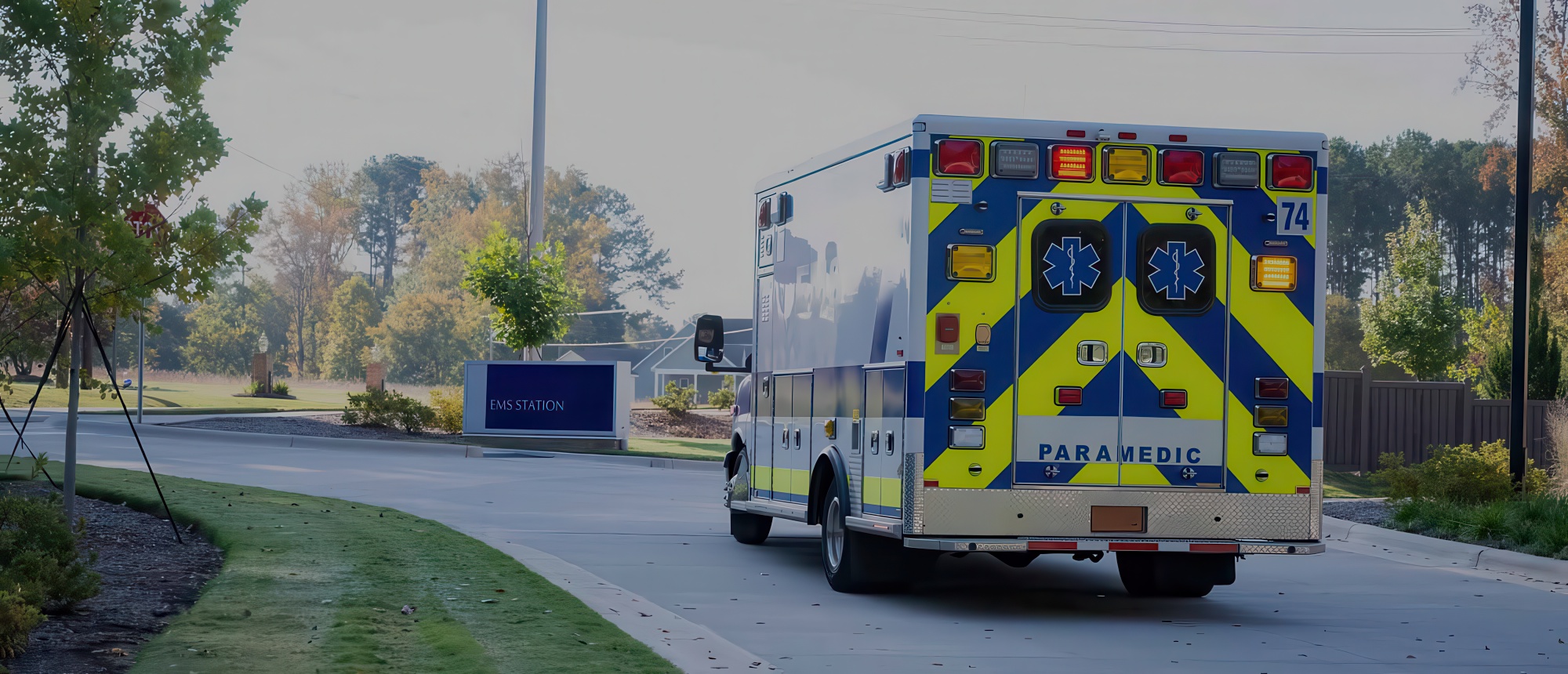A paramedic ambulance with yellow and blue chevron markings drives past an EMS station sign on a quiet suburban road.