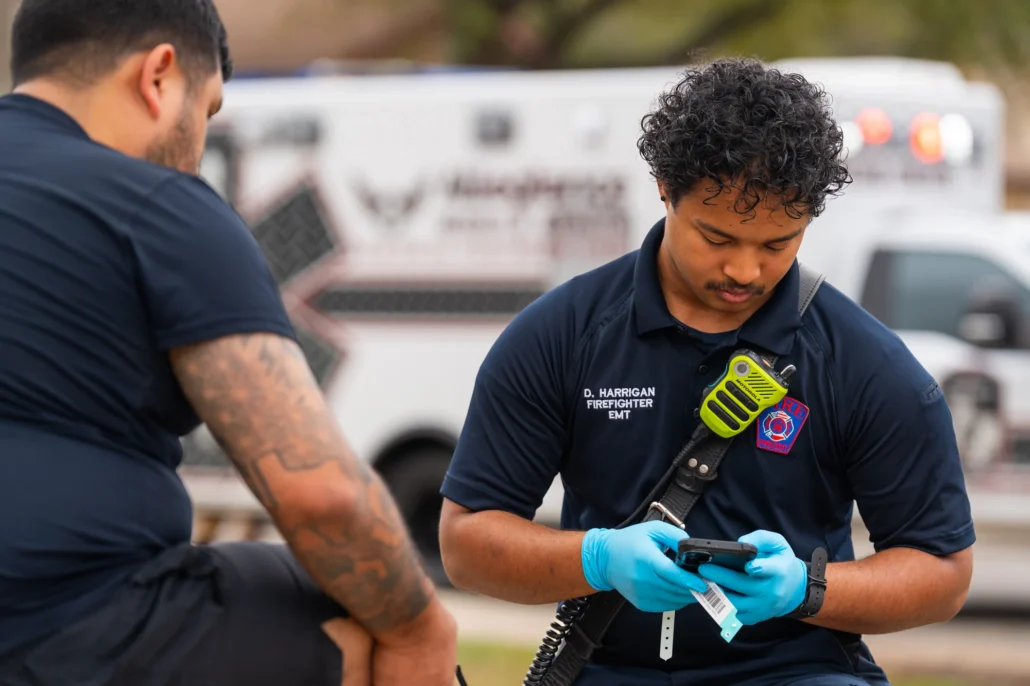 A firefighter in uniform and blue gloves reviews patient information on a smartphone while seated outdoors, with an ambulance visible in the background.