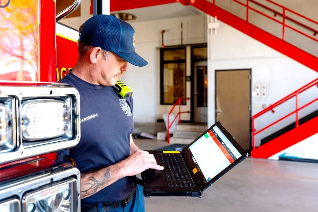 A paramedic with a tattooed arm and a blue cap stands beside a fire truck, using a laptop inside a fire station.