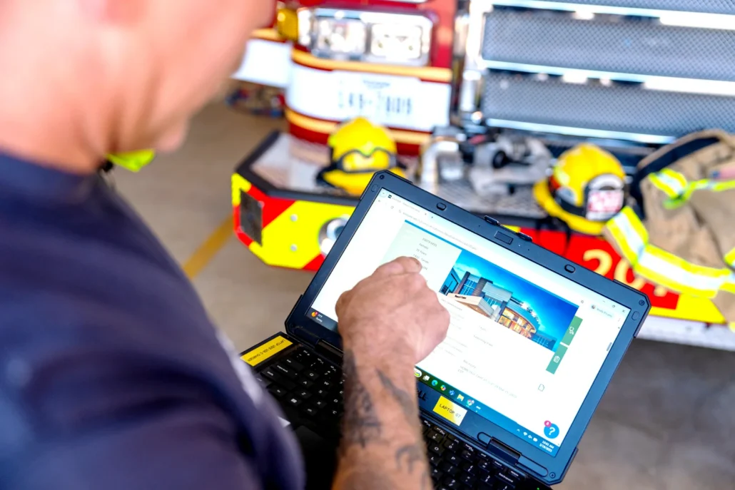 A firefighter with a tattooed arm uses a laptop to access a digital form while seated in a fire station, with a red fire truck and yellow helmets visible in the background.