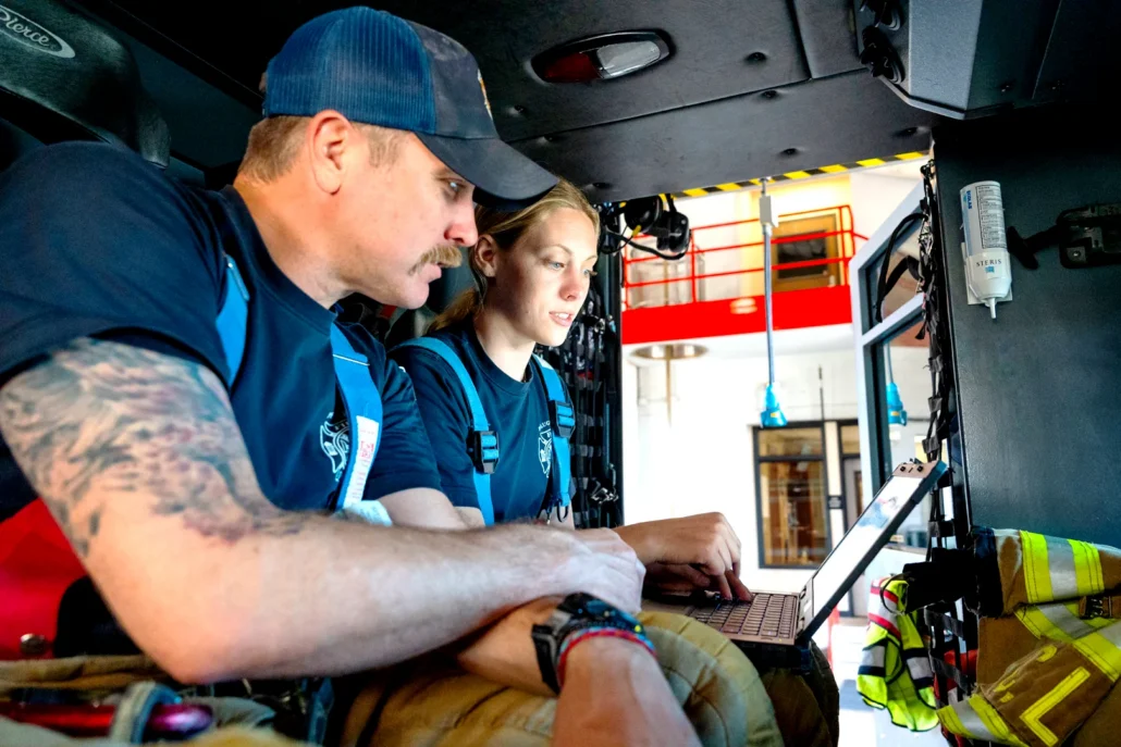 Two emergency responders in blue uniforms sit inside a fire vehicle, reviewing data on a laptop with fire gear visible nearby.