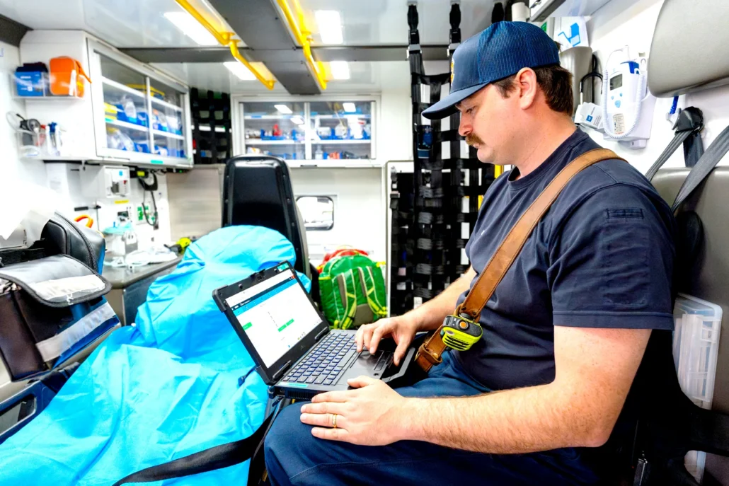 A first responder in a blue uniform and cap sits inside an ambulance, typing on a laptop while attending to a patient covered by a blue blanket.