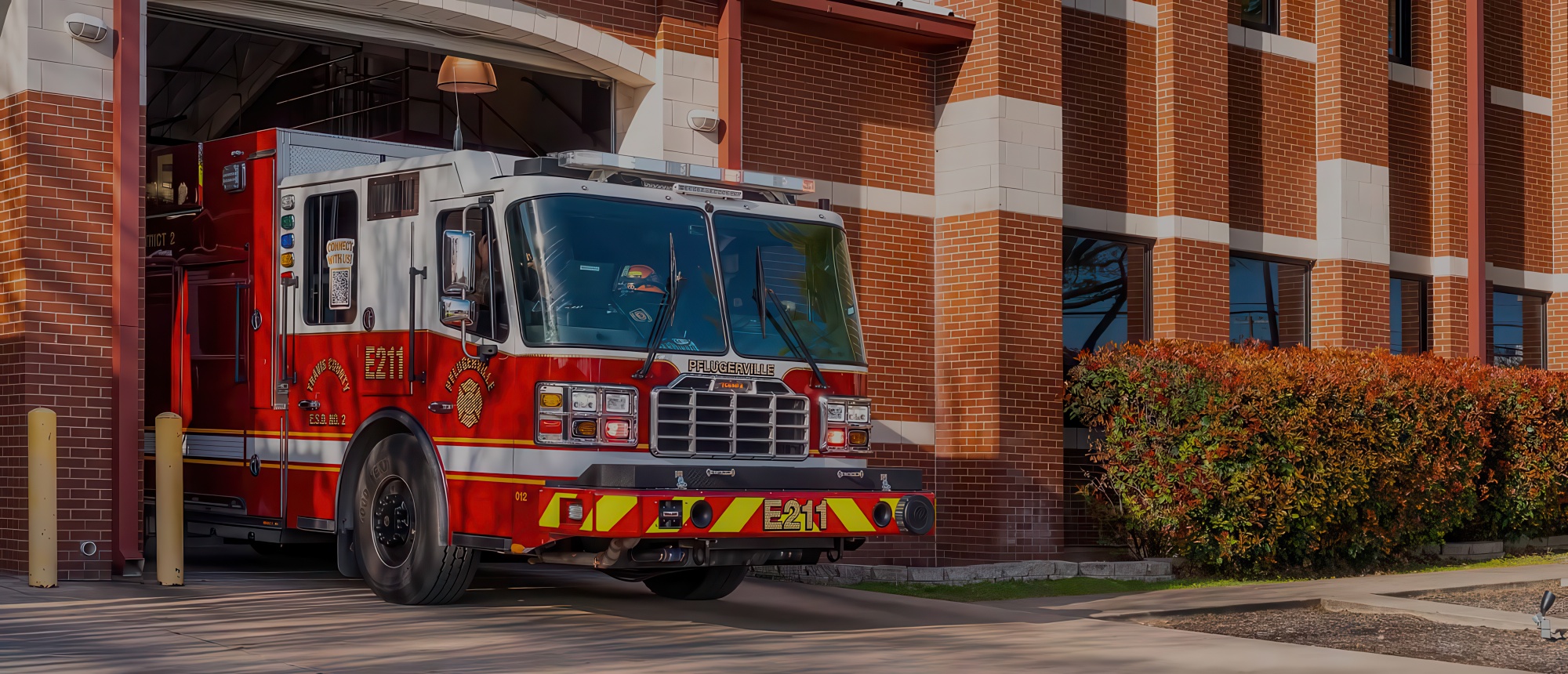 A red and white fire truck, is parked partially inside its brick fire station bay with the garage door open.
