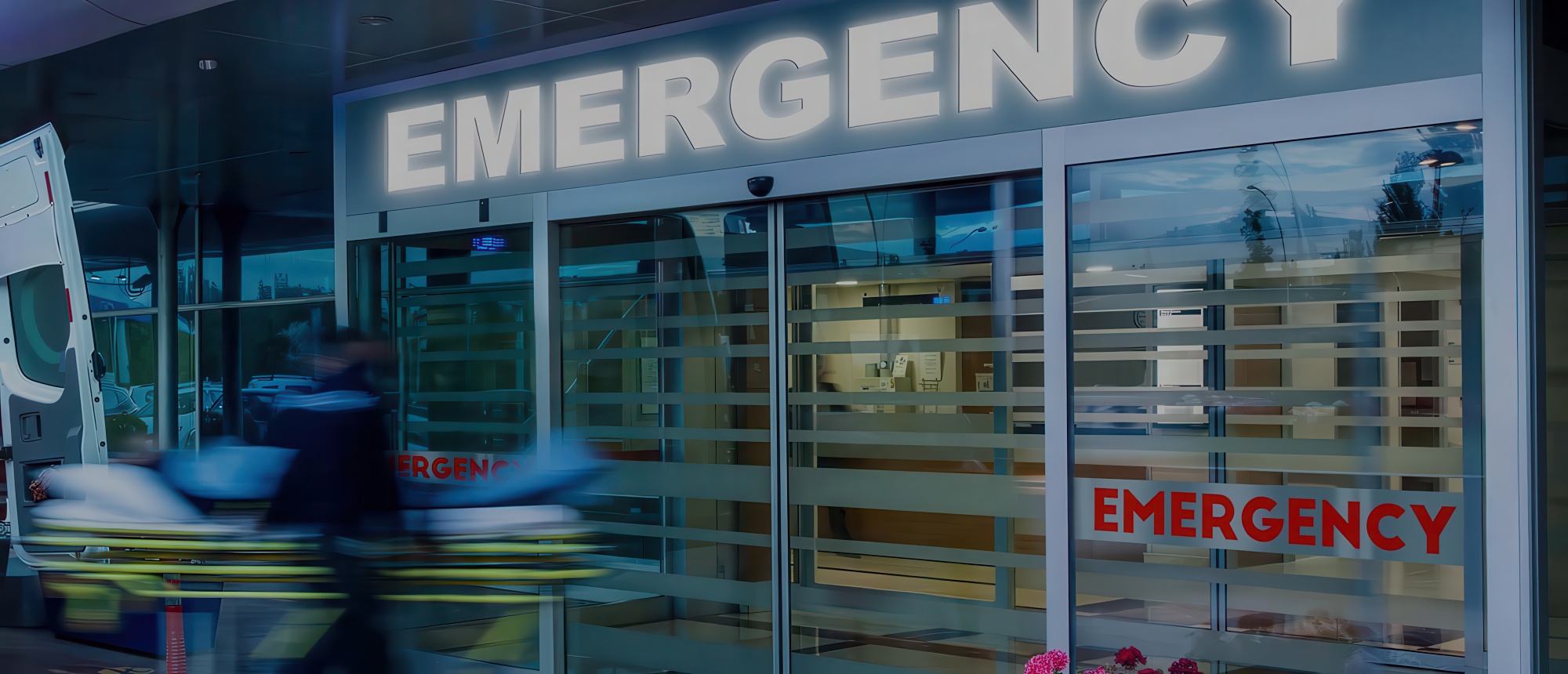 Emergency room entrance with illuminated "EMERGENCY" sign above glass doors, medical staff moving a gurney outside an ambulance at dusk.