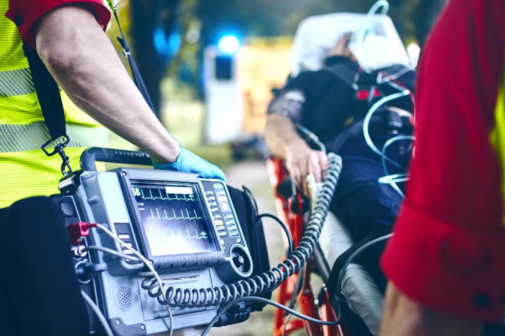 A paramedic in a high-visibility vest holds a cardiac monitor displaying vital signs while attending to a patient on a stretcher outdoors.