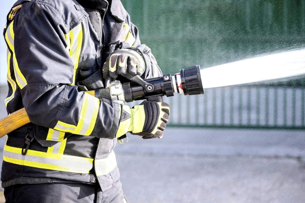 A firefighter in a turnout coat with reflective stripes holds a hose nozzle spraying a strong stream of water against a blurred green background.