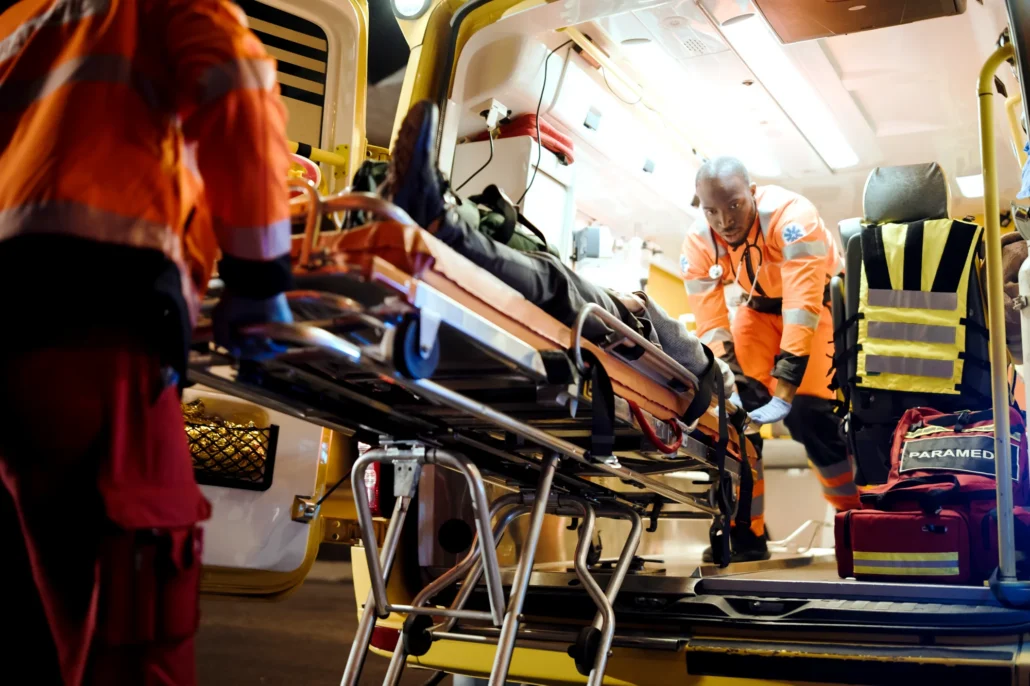 Paramedics in orange high-visibility uniforms attend to a patient on a gurney inside an ambulance.