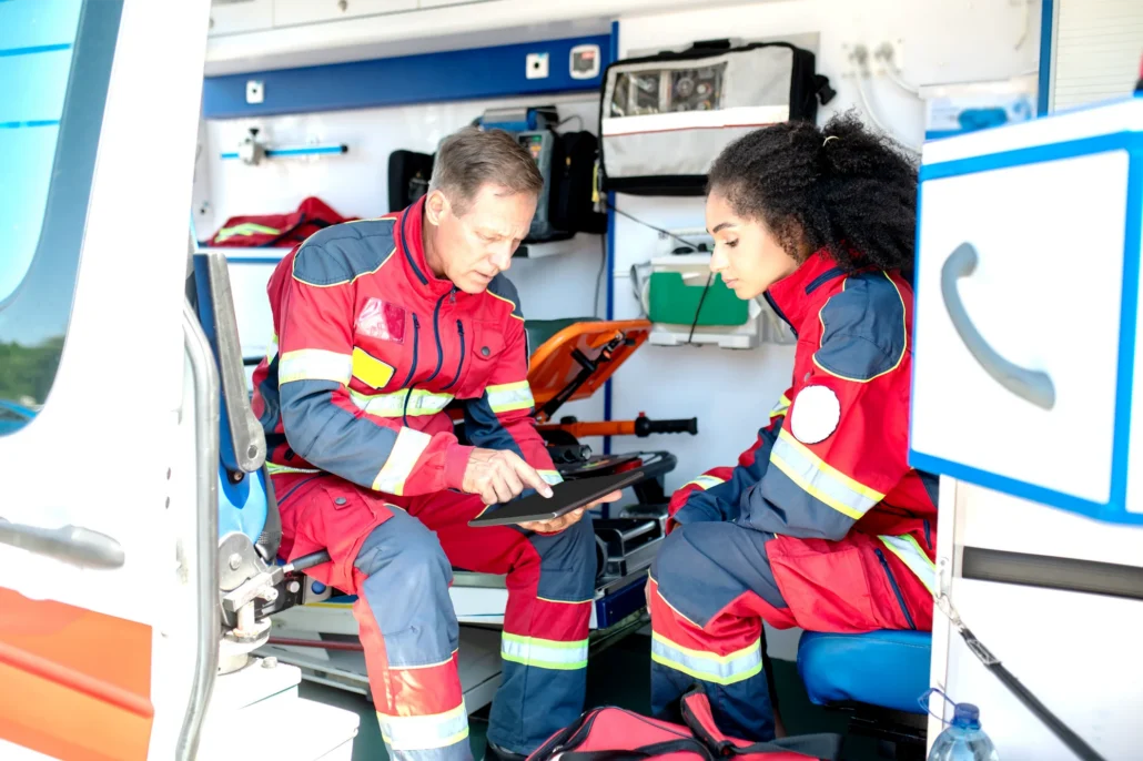 Two paramedics in red and blue uniforms review patient information on a tablet inside the back of an ambulance.