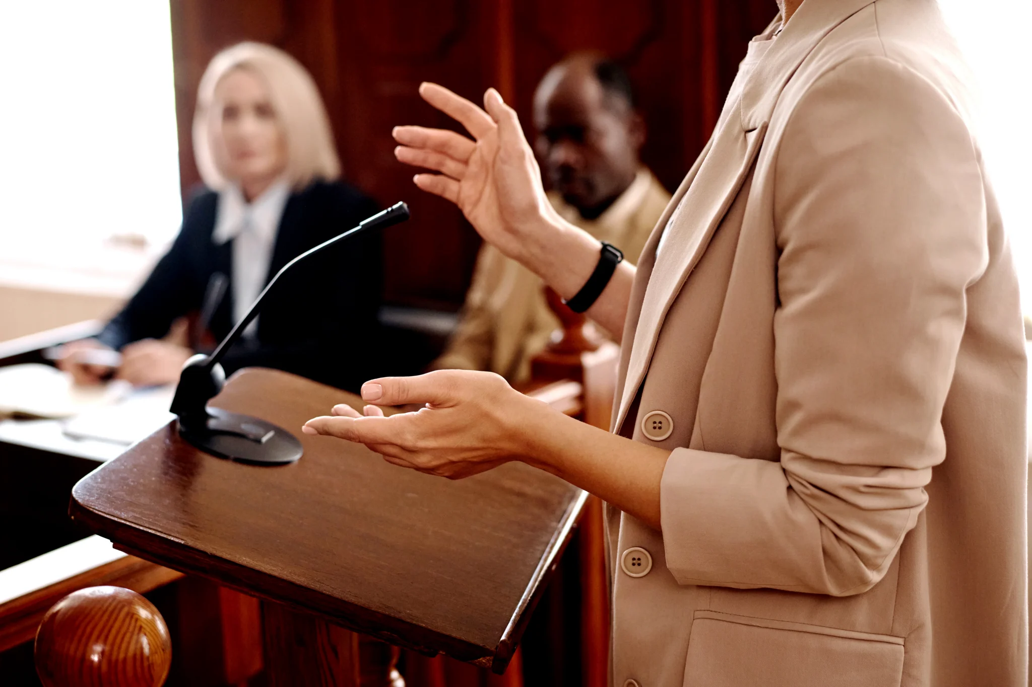 A lawyer in a beige suit gestures while speaking at a wooden podium in a courtroom, with the judge and jury visible in the background.