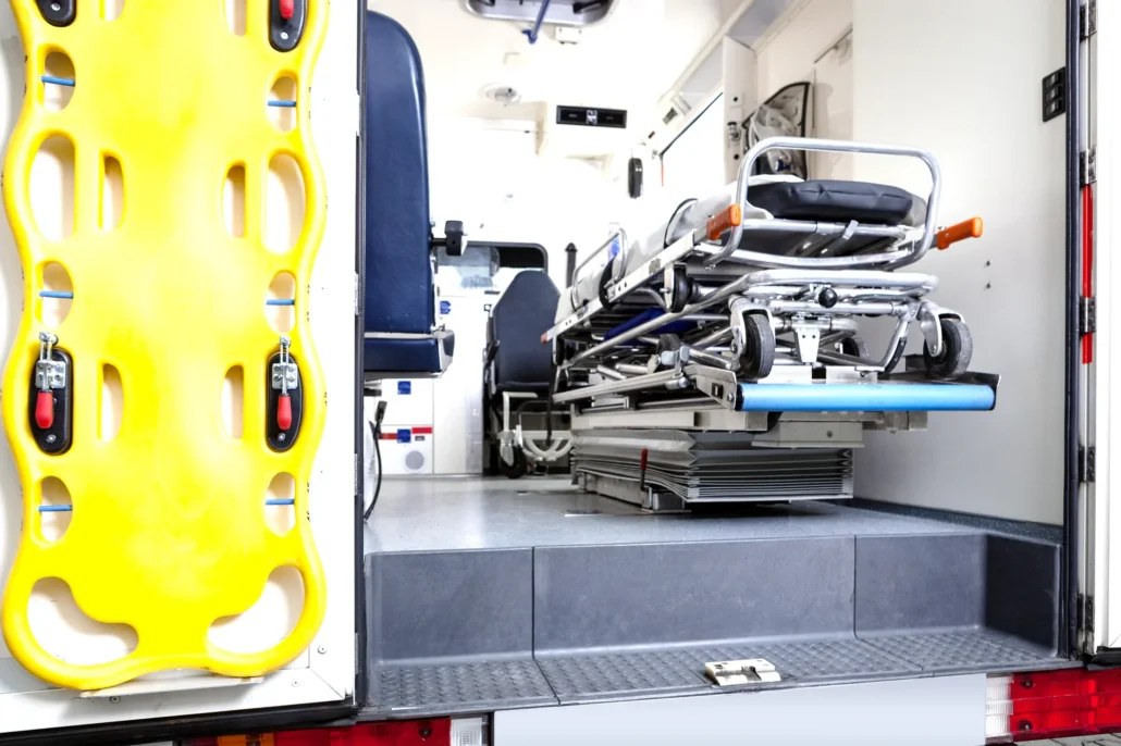 The interior of an ambulance features a yellow spine board mounted on the left wall and a stretcher with a black mattress positioned in the center.