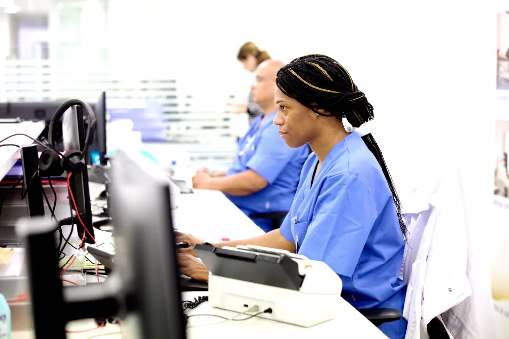 A female nurse in blue scrubs with braided hair sits at a workstation, typing on a computer keyboard in a busy hospital setting.