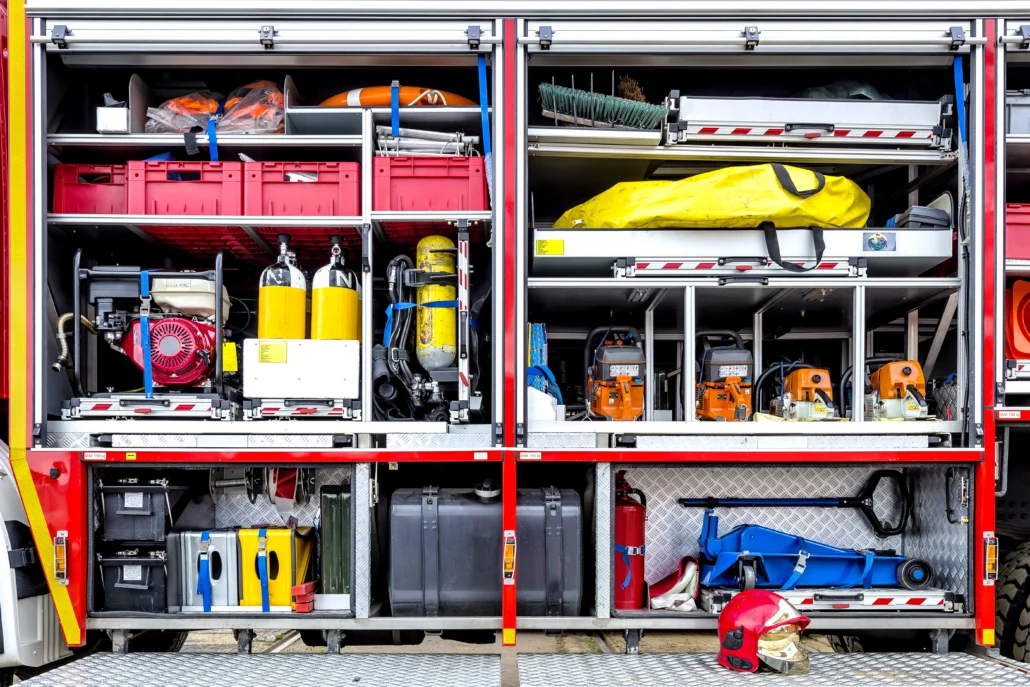 The open side compartments of a fire truck reveal organized equipment including red storage bins, yellow oxygen tanks, a red generator, orange power tools, and a red helmet resting on the floor.