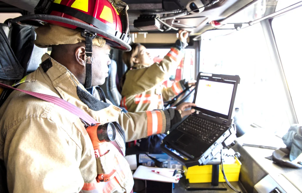 A firefighter in full gear sits inside a fire truck, using a laptop mounted on the dashboard while his partner reaches for equipment above.